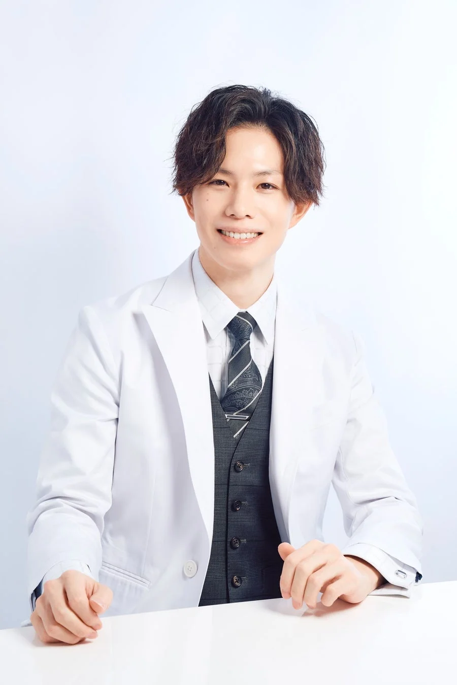白い白衣を着用した若い日本人男性が、笑顔でカメラを見つめています。