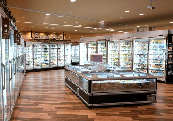 スーパーマーケットの冷凍食品売り場です。ガラス扉の冷凍ケースや、中央に低い冷凍ケースが並び、様々な冷凍食品が陳列されています。店内は明るく、床は木目調です。