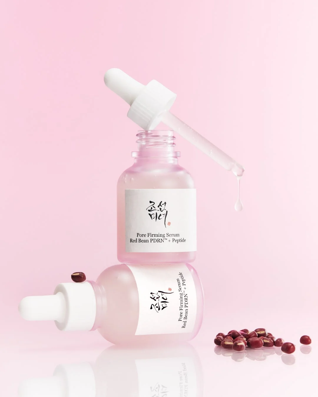 ピンクの背景に、小豆を配合した美容液のボトルが2本配置されています。1本は立ててあり、もう1本は横たわっており、スポイトから滴が垂れています。