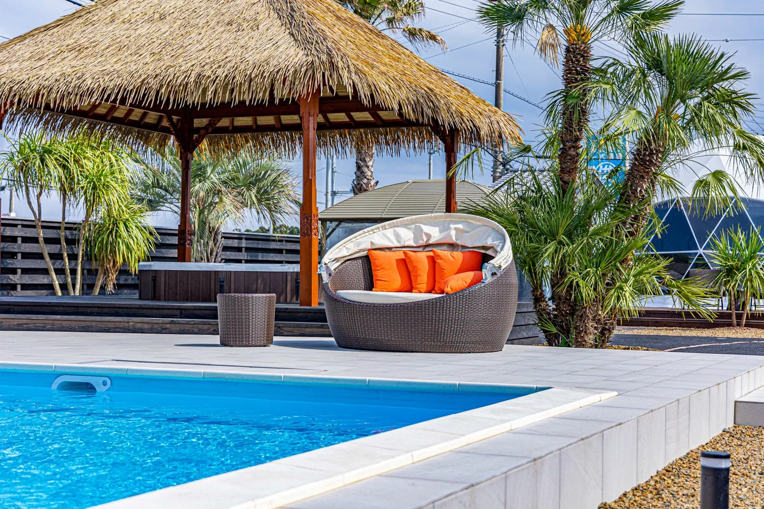 青いプールと藁葺き屋根のガゼボ、デイベッドが配置されたリゾート施設