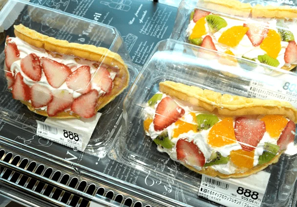 透明なプラスチック容器に入った、いちごや様々なフルーツとホイップクリームがたっぷり挟まれたワッフルサンドが複数並べられています。価格表示も見えます。