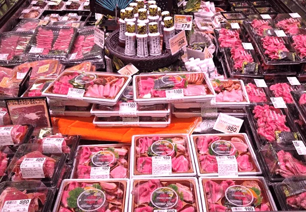 スーパーマーケットの精肉売り場が写されています。牛肉や豚肉のスライス、ローストビーフのような加工肉など、多種多様なパック詰めされた肉が冷蔵ケースにぎっしりと並べられており、多くの商品に価格が表示されています。