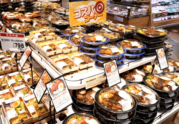 スーパーマーケットの惣菜コーナーで、様々な種類の弁当や調理済み食品が大量に陳列されています。「コスパコーナー」と書かれたポップが目を引き、手軽に購入できる食事が並んでいます。