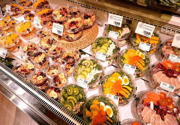 スーパーマーケットの惣菜コーナーで、色とりどりのサラダやフルーツグラノーラが多数陳列されています。様々な食材を使った健康的なデリ商品が、透明な容器に入って販売されており、価格も表示されています。