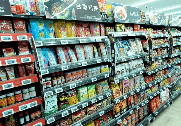 スーパーマーケットの食品売り場が写されており、台湾料理、麻辣湯、ラーメンなどのアジア系パッケージ食品や調味料が棚にぎっしり並んでいます。多くの商品に価格表示が見られます。