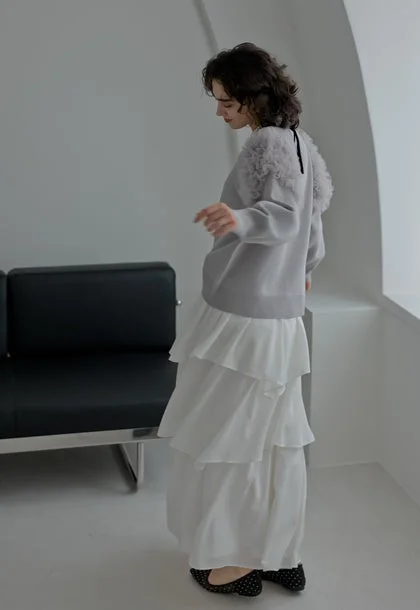 ふわふわの飾りがついたグレーのセーターと白いティアードロングスカートを着用した女性