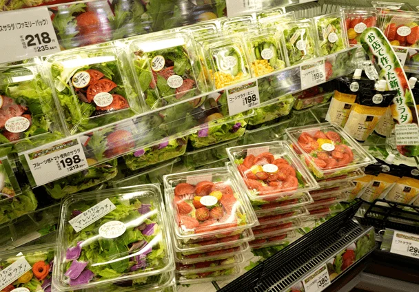 スーパーマーケットの冷蔵ケースに陳列された、様々な種類のパック入りサラダの様子です。レタス、トマト、コーンなどが入ったサラダが並び、価格表示やプロモーションのPOPも見えます。