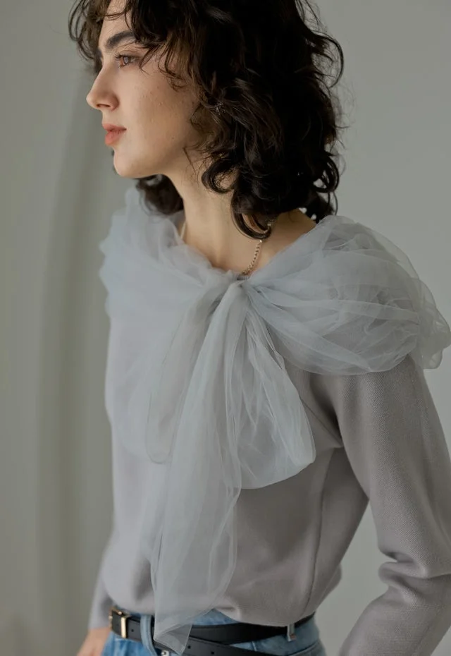 横顔の女性が、グレーのニットに同色のチュールをあしらったトップスを着て、自然な雰囲気で立っているポートレート