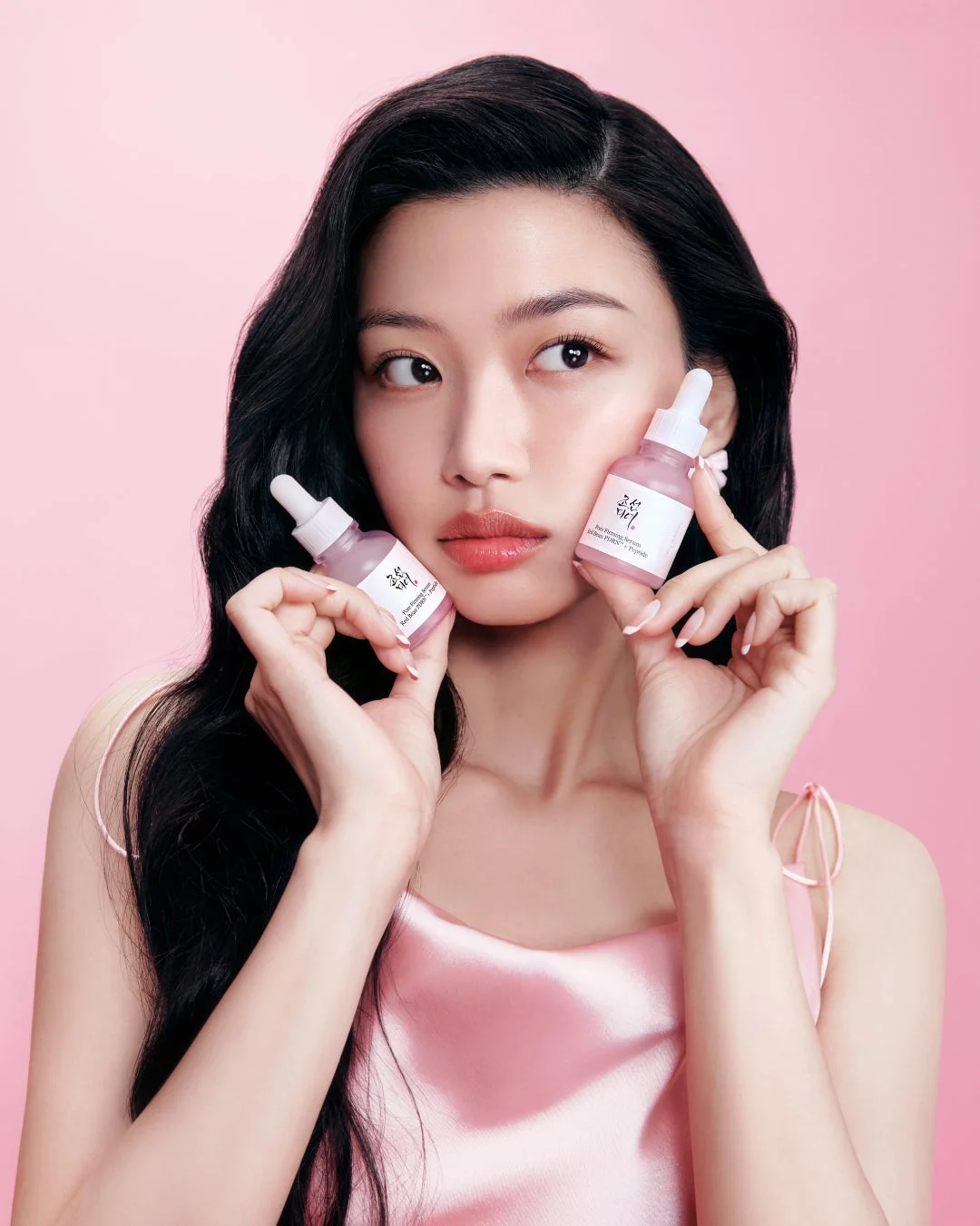 ピンクの背景とサテンのトップスを着用した女性が、2本のピンク色の美容液ボトルを手に持っています。