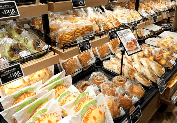 スーパーマーケットのパン売り場で、個包装されたメロンパン、あんぱん、マフィン、シナモンロールなど、様々な種類の菓子パンが豊富に陳列されています。価格表示も見られます。