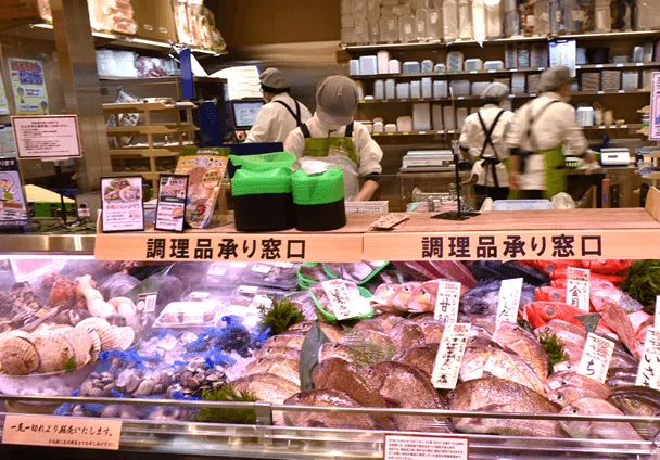 日本の鮮魚売り場の様子。新鮮な魚や貝類が氷のショーケースに並べられ、「調理品承り窓口」の看板が見える。数名の店員が作業しており、購入した魚の調理サービスも提供している様子。