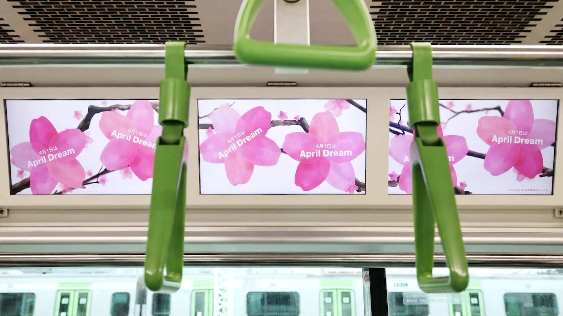 電車内で撮影された画像で、上部のデジタルサイネージにはピンク色の桜の花と「4月1日は April Dream」という広告が表示されています。