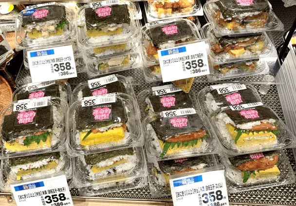 スーパーマーケットの陳列棚に並べられた、おにぎりと弁当の中間のような「おにぎらず」が写っています。鶏マヨ、ツナマヨ、厚焼玉子、スパムなどの様々な具材が海苔とご飯で挟まれており、「運命的相性!」というキャッチフレーズと共に新商品として358円で販売されています。