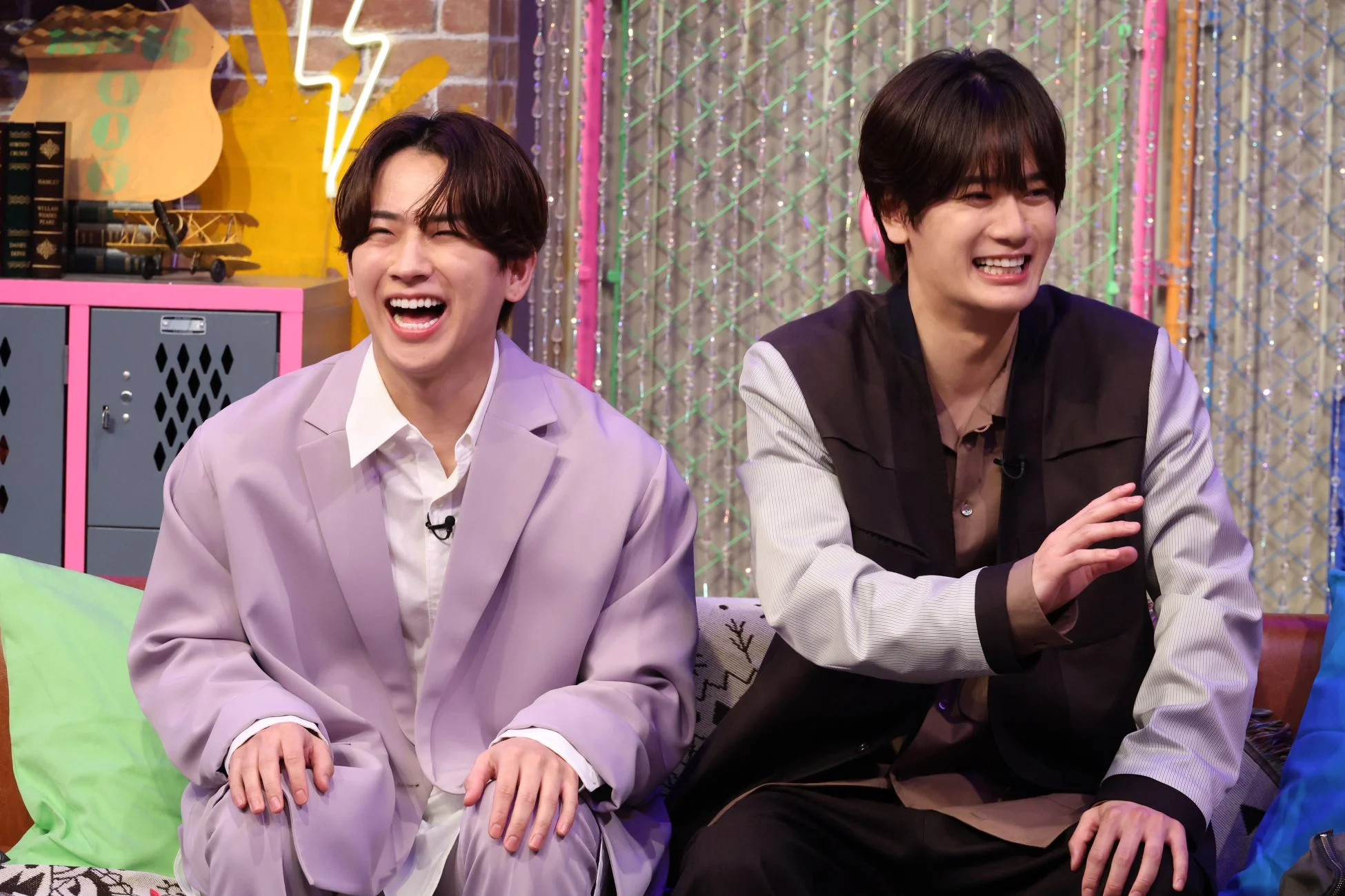 2人の若い男性がスタジオらしき場所で、とても楽しそうに笑っている瞬間を捉えた画像です。カラフルな背景の前で、彼らはリラックスした表情を見せており、和やかな雰囲気が伝わってきます。