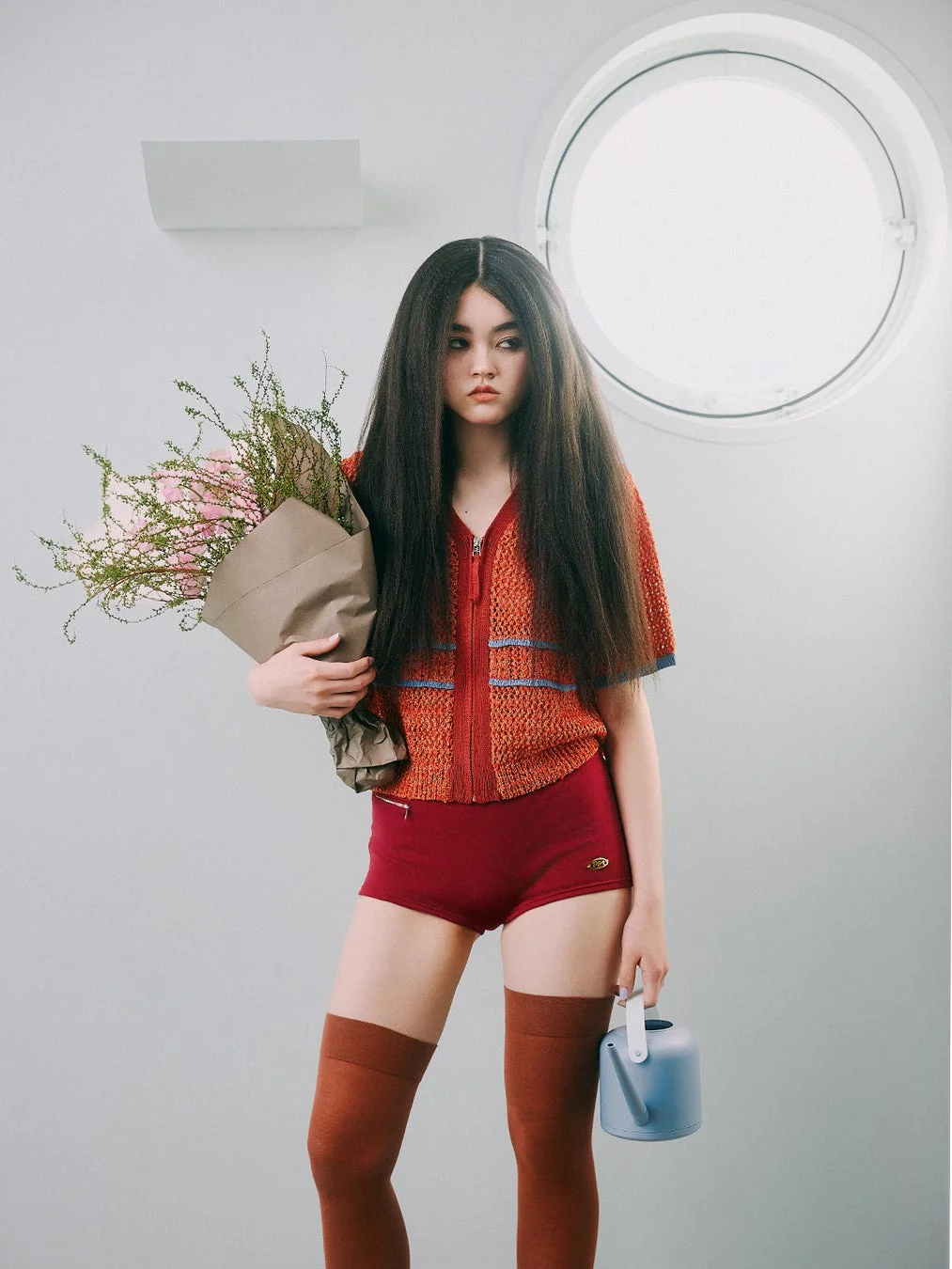 長い黒髪の女性が花束とじょうろを持ち、赤いショートパンツとオレンジ色のニットを着て立つポートレート。