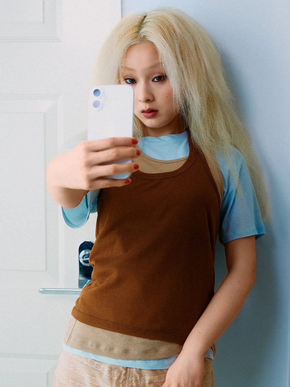 明るいブロンドの長いカーリーヘアの若い女性が、白いスマートフォンを持ってセルフィーを撮るようなポーズをとっています。