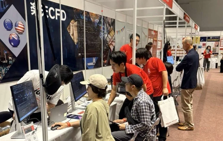 展示会のような会場で、複数の人々がブースに設置されたコンピュータの画面を見ている様子。