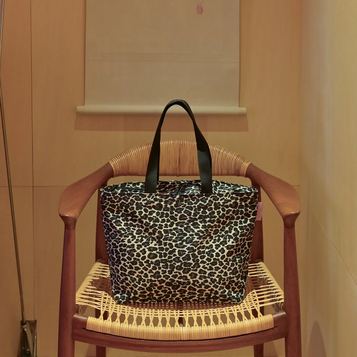 ヒョウ柄のトートバッグが、籐編みの座面を持つ木製の椅子に置かれています。暖色系の壁を背景に、室内の落ち着いた雰囲気が感じられます。