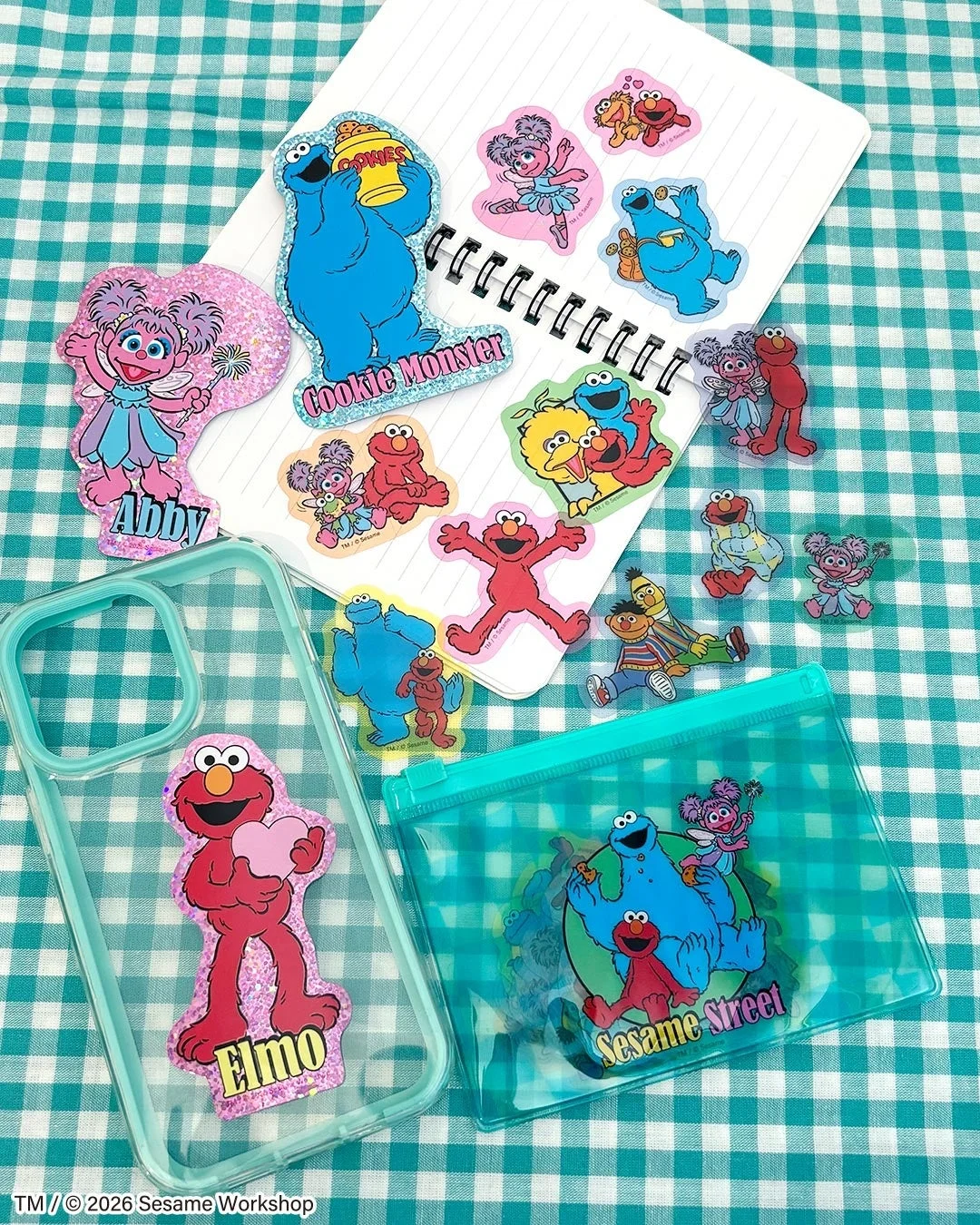セサミストリートのキャラクターたちがデザインされた、様々な種類のステッカーが散りばめられています。ノート、スマートフォンケース、クリアポーチなどのアイテムも一緒に写っており、可愛らしい商品展開が伺える画像です。