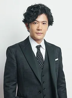 濃い色のスリーピーススーツを着用し、水玉模様のネクタイを締めた男性のポートレートです。カメラをまっすぐ見ており、特徴的な巻き毛の髪型をしています。