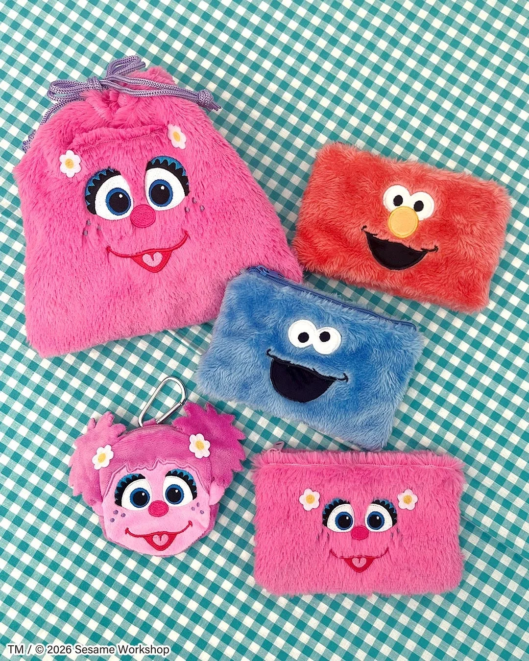 セサミストリートのキャラクター、アビー・カダビー、エルモ、クッキーモンスターをモチーフにしたふわふわのポーチや巾着が、緑と白のギンガムチェックの布の上に並べられています。可愛らしいデザインの小物入れです。