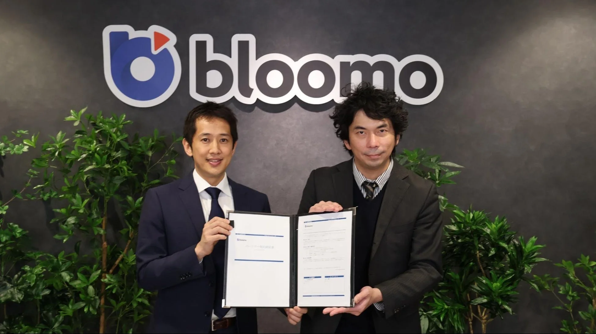 bloomoのロゴが背景にある部屋で、2人の男性が笑顔で契約書を掲げている画像