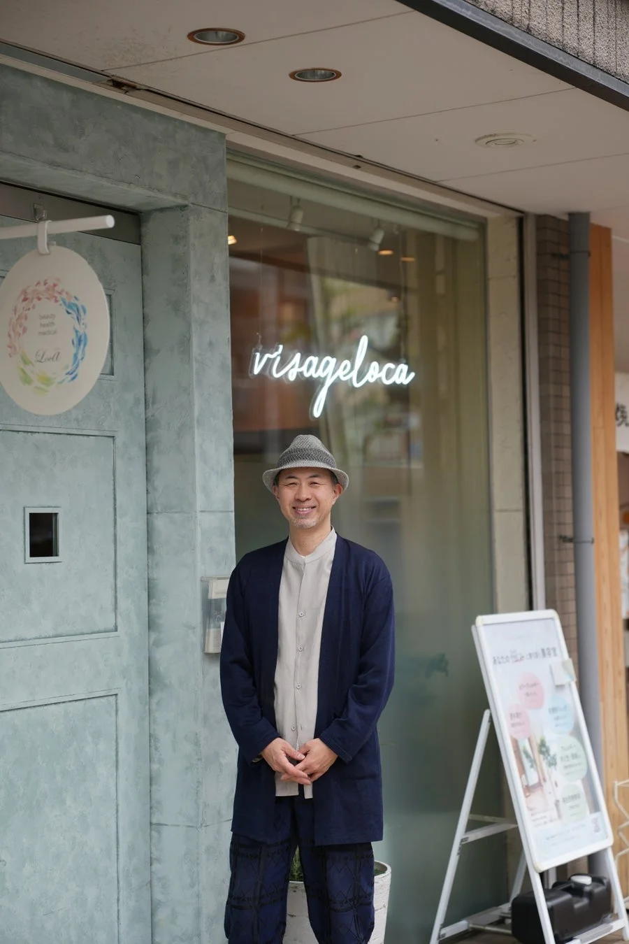 グレーの帽子をかぶり、ネイビーのジャケットとライトカラーのシャツを着た男性が、店舗の前に立って笑顔でカメラを見つめている。