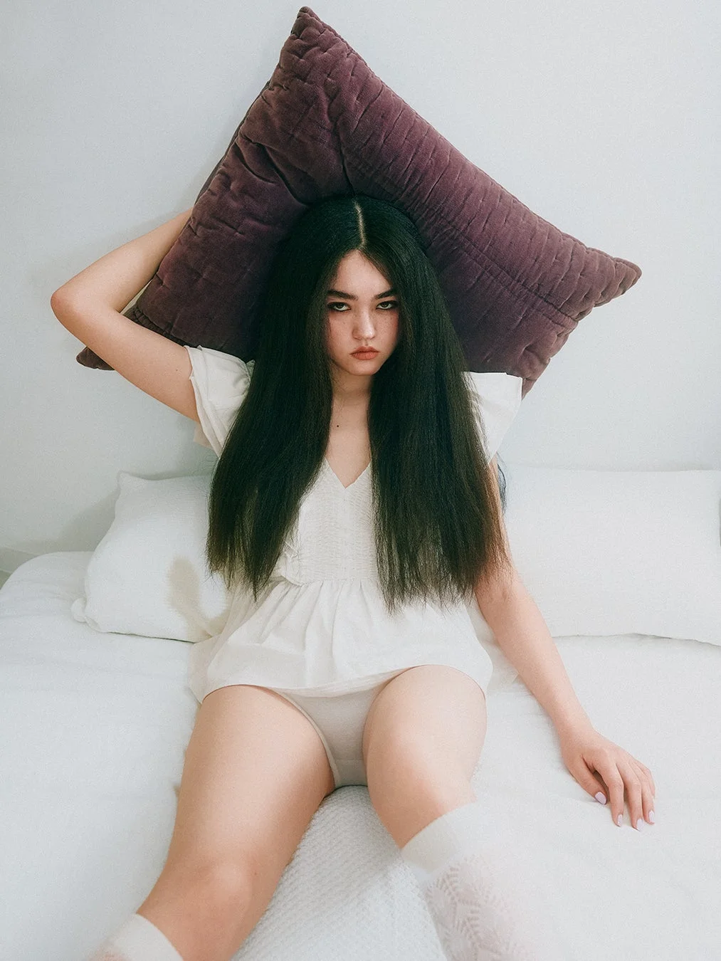 長い黒髪の女性が白いベッドに座り、頭上に大きな紫色の枕を抱えています。