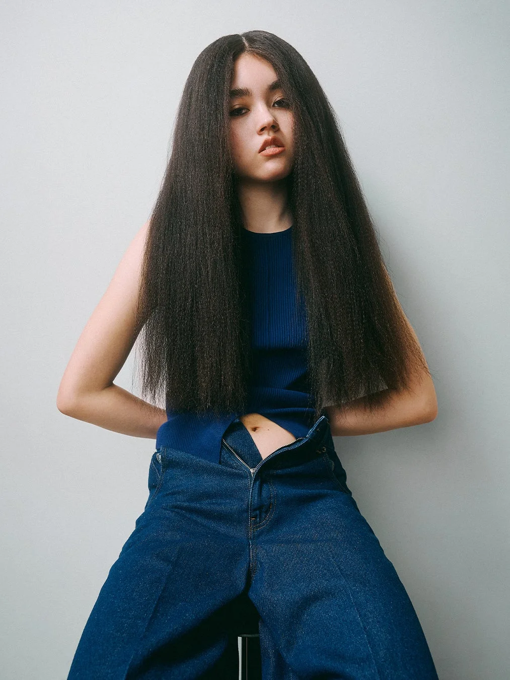 長い黒髪の女性が青いトップスとデニムを着用し、ジッパーを下ろした状態で座っているポートレート。