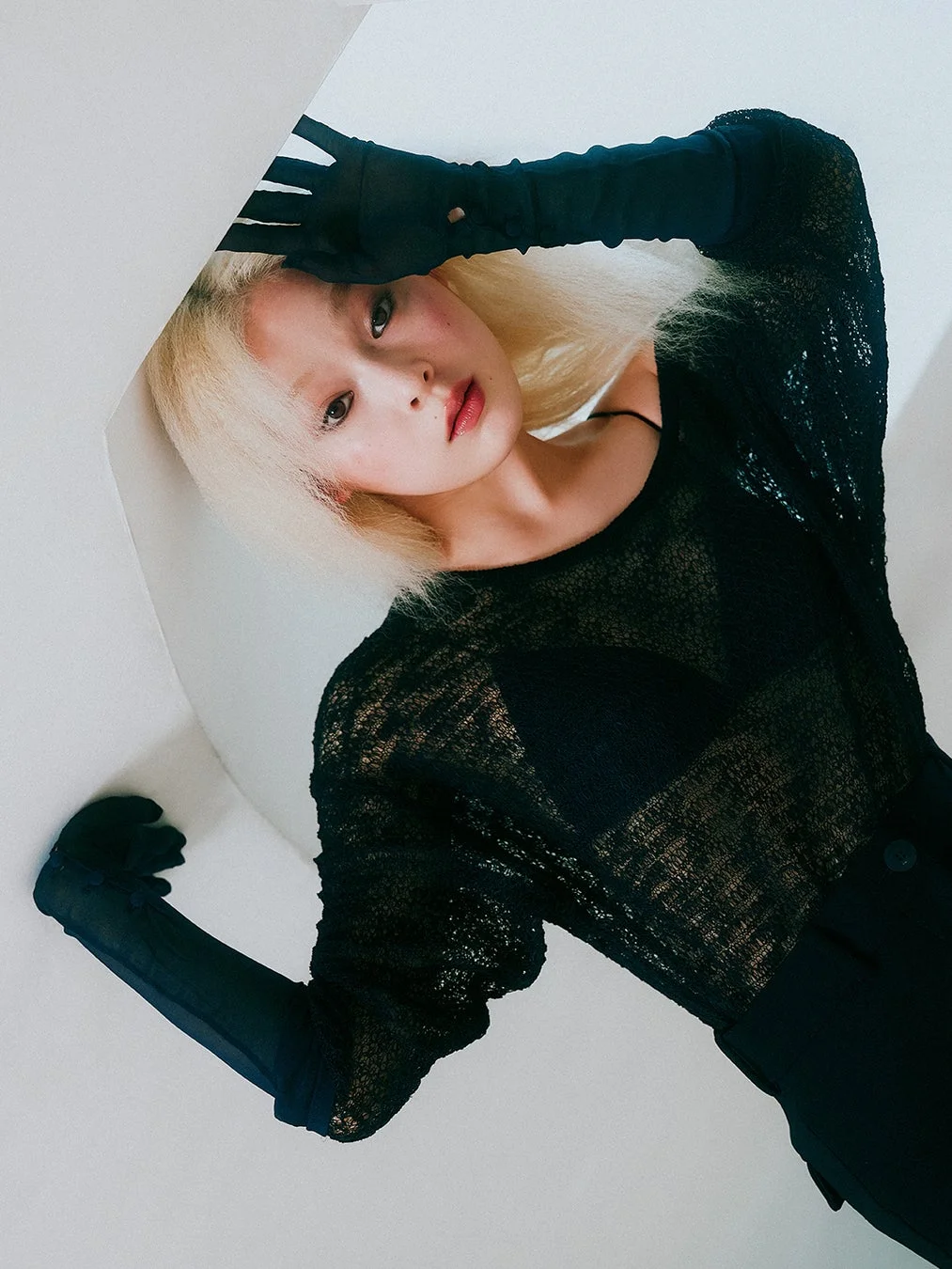 白い背景の前で、ブロンドヘアの女性が黒いシースルートップとロンググローブを着用し、腕を上げてポーズをとっている。