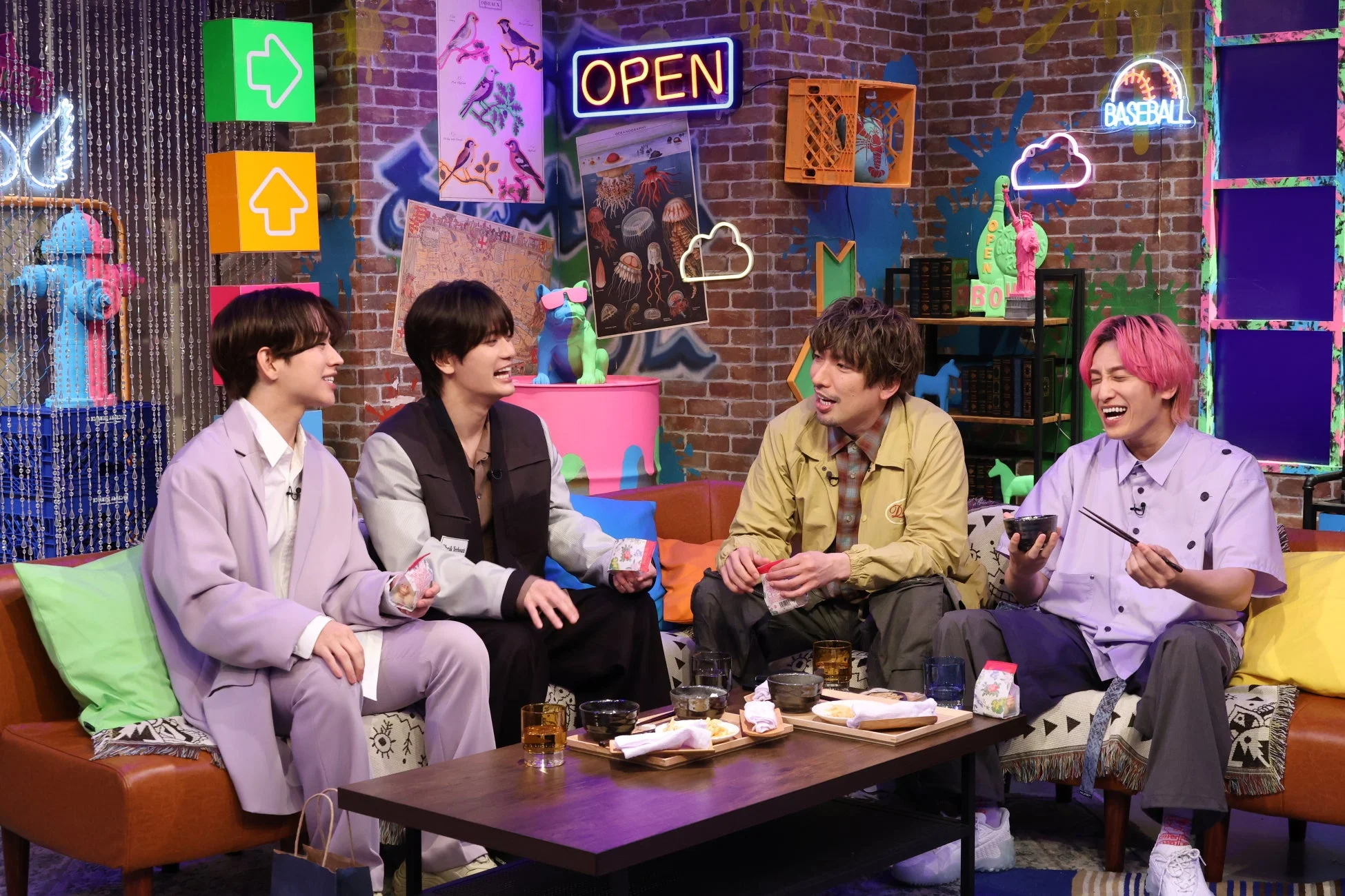 カラフルなセットのスタジオで、4人の男性がソファに座り、食事をしながら楽しげに談笑している様子です。ポップなネオンサインや装飾が特徴的な空間で、リラックスした雰囲気が伝わってきます。