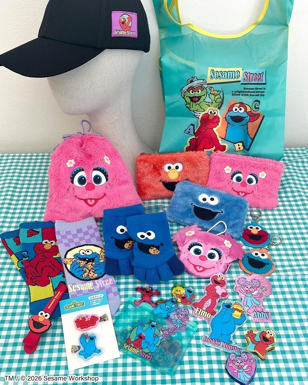 セサミストリートのカラフルなキャラクターグッズが多数集められた写真です。エルモ、クッキーモンスター、アビーカダビーなどがデザインされた帽子、エコバッグ、ポーチ、靴下、手袋、ヘアクリップなどがチェック柄のテーブルに並んでいます。