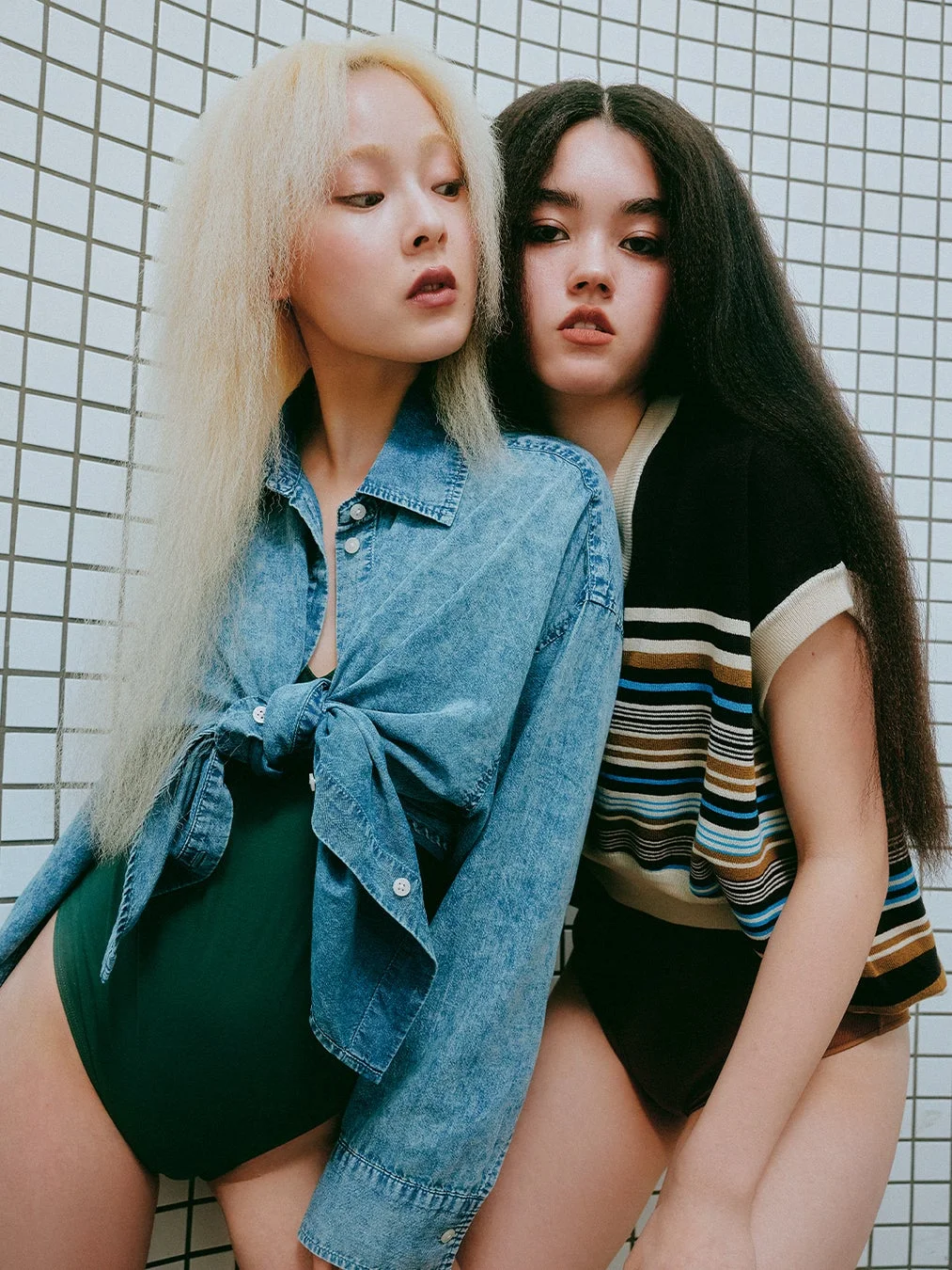 個性的なヘアスタイルの二人の女性が、デニムシャツやストライプのトップスを着用し、白いタイル壁を背景にポーズをとっています。