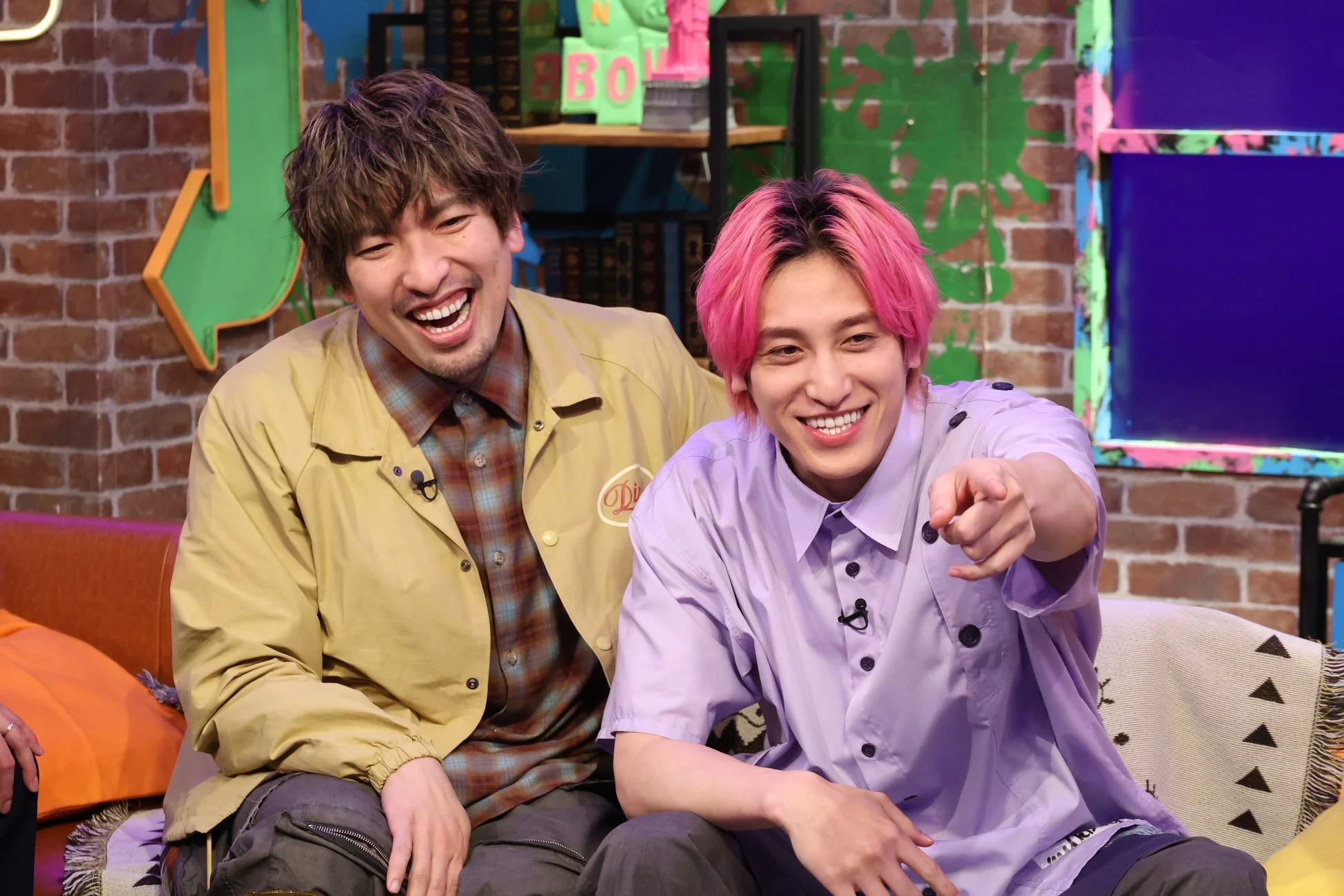 カラフルなセットで笑顔を見せる二人の男性。一人はピンク髪で指を指しており、もう一人は茶髪で大きく笑っています。楽しそうな雰囲気の場面です。