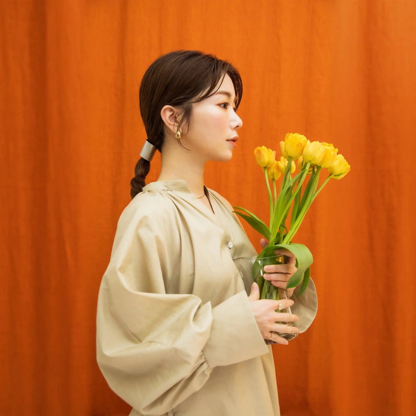 オレンジ色の背景を背に、横顔の女性が黄色のチューリップが入った花瓶を抱えています。