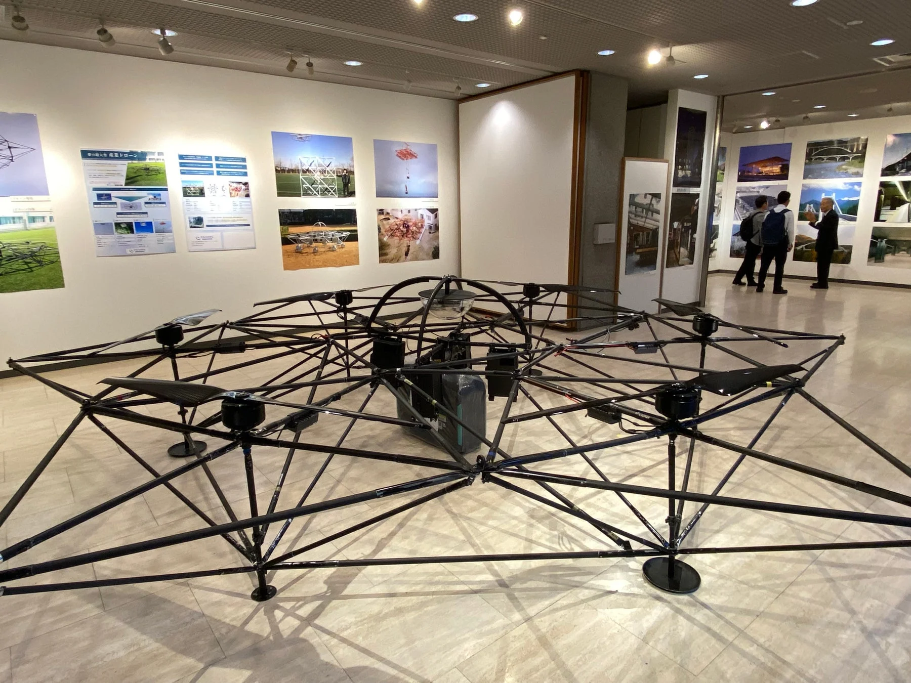 広い展示空間に大型の産業用ドローンの骨組みが展示されています。壁にはドローンの用途や技術を紹介するポスターが貼られ、来場者が他の展示物を見ている様子が写っています。
