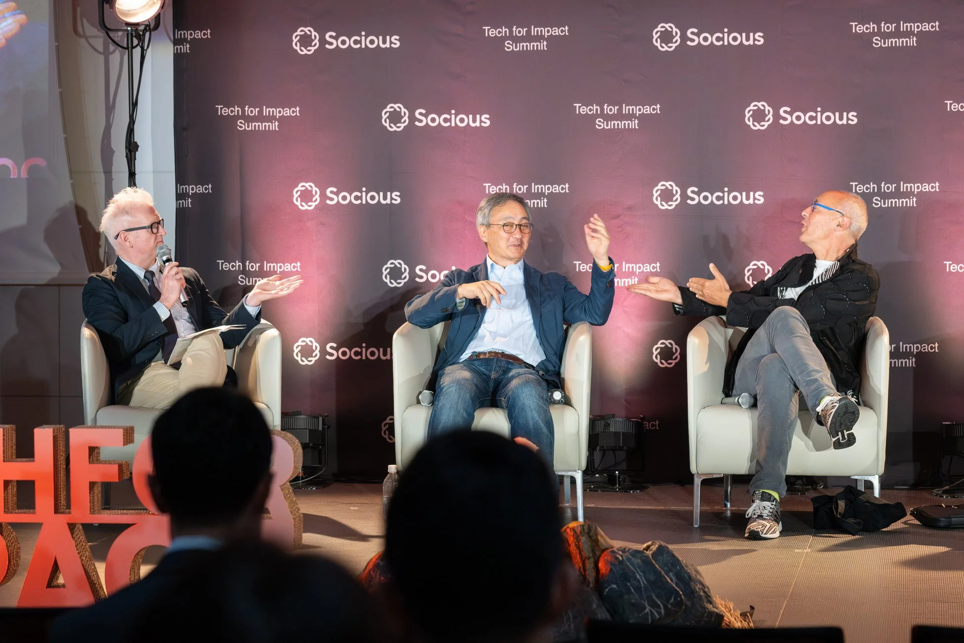 Tech for Impact Summitと書かれた背景の前で、3人の男性がステージ上でパネルディスカッションを行っている様子。