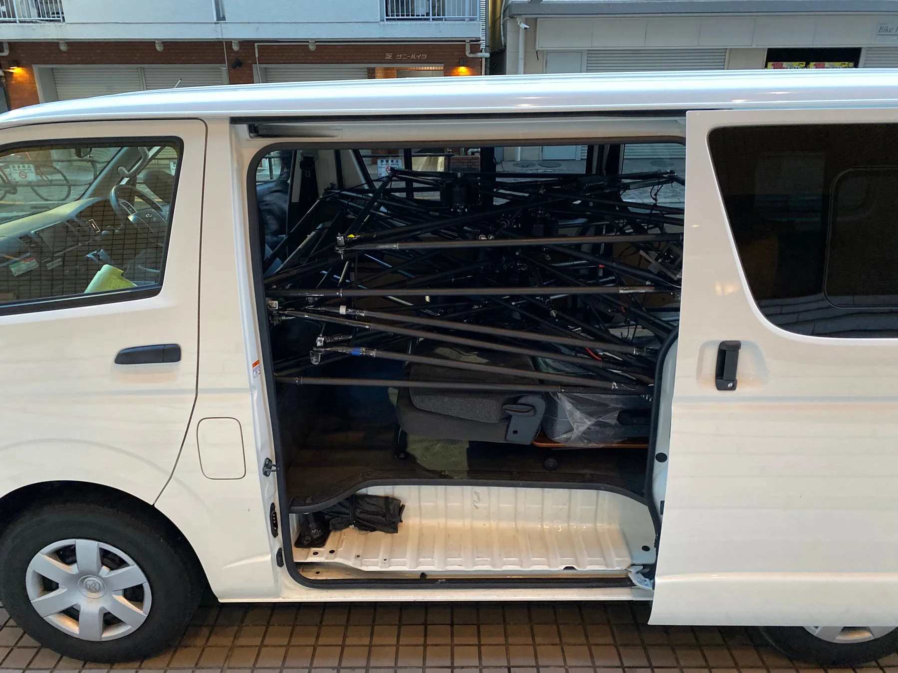 白いワンボックスカーの側面ドアが開いており、車内には多数の黒い金属製の棒が積み込まれています。車両は建物の横に駐車されており、ビジネス用途の荷物運搬の様子がうかがえます。