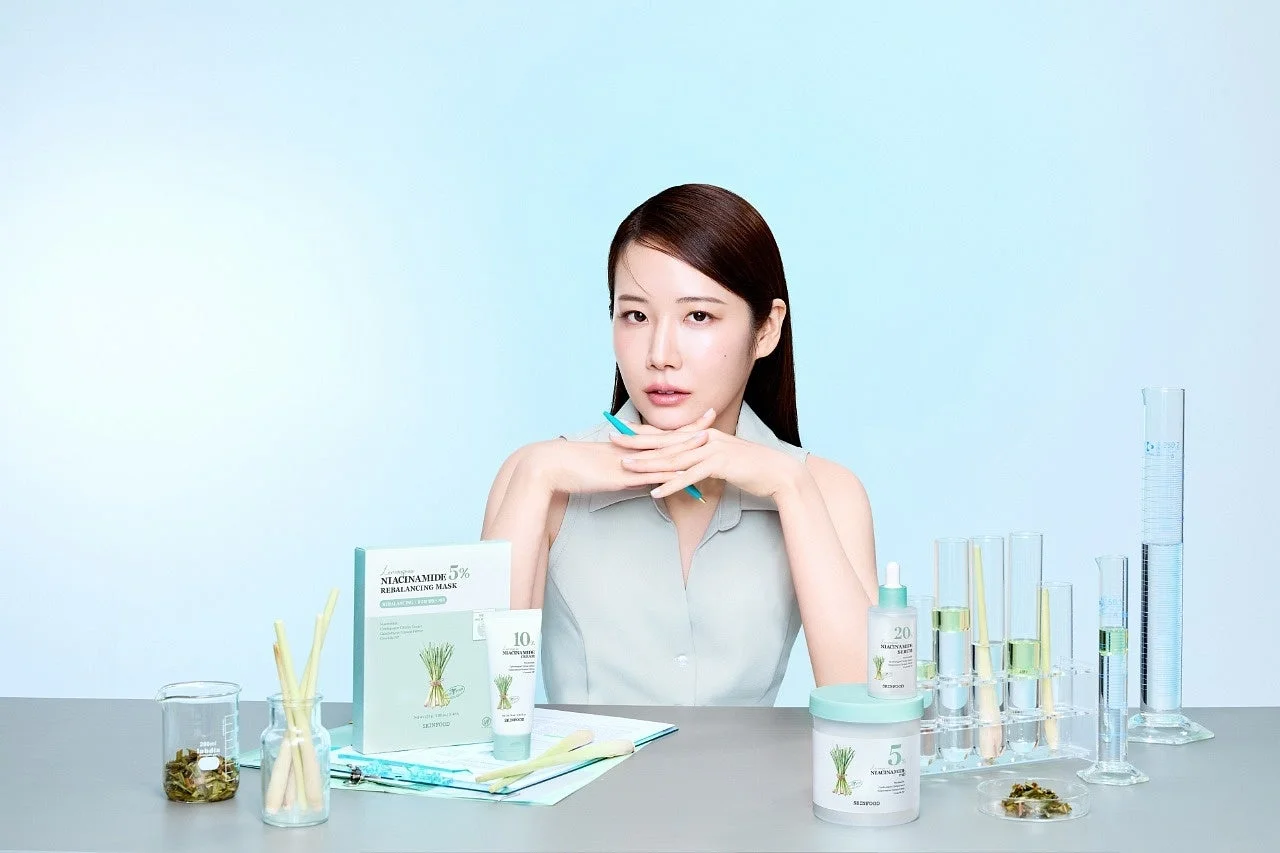 ナイアシンアミド配合のスキンケア製品と実験器具が並ぶテーブルの前に、モデルの女性が座っています。清潔感のある背景で、科学と自然を融合した美容製品のイメージを伝えています。