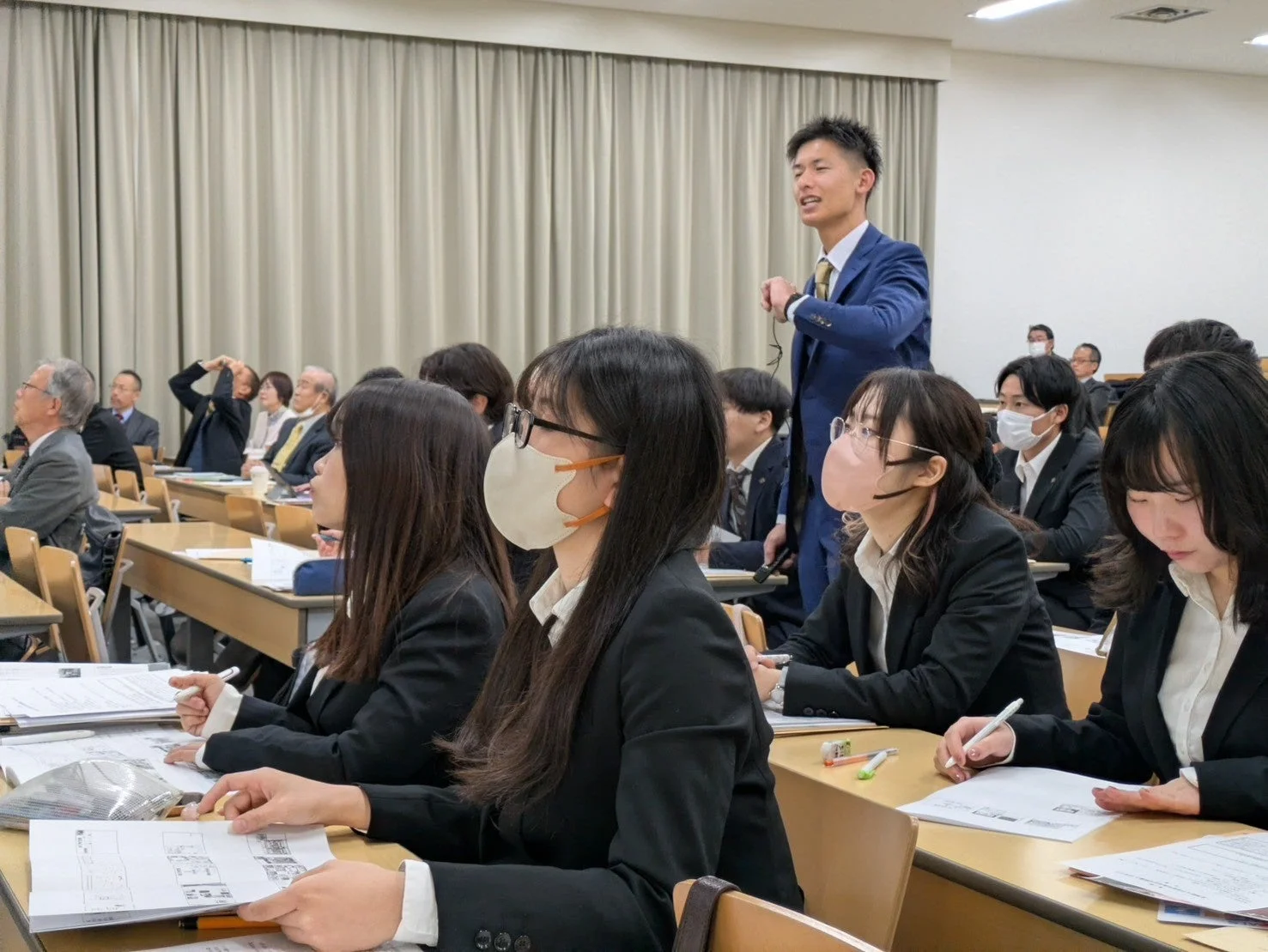 教室のような空間で、スーツ姿の参加者たちが講義やセミナーに真剣に取り組んでいる様子