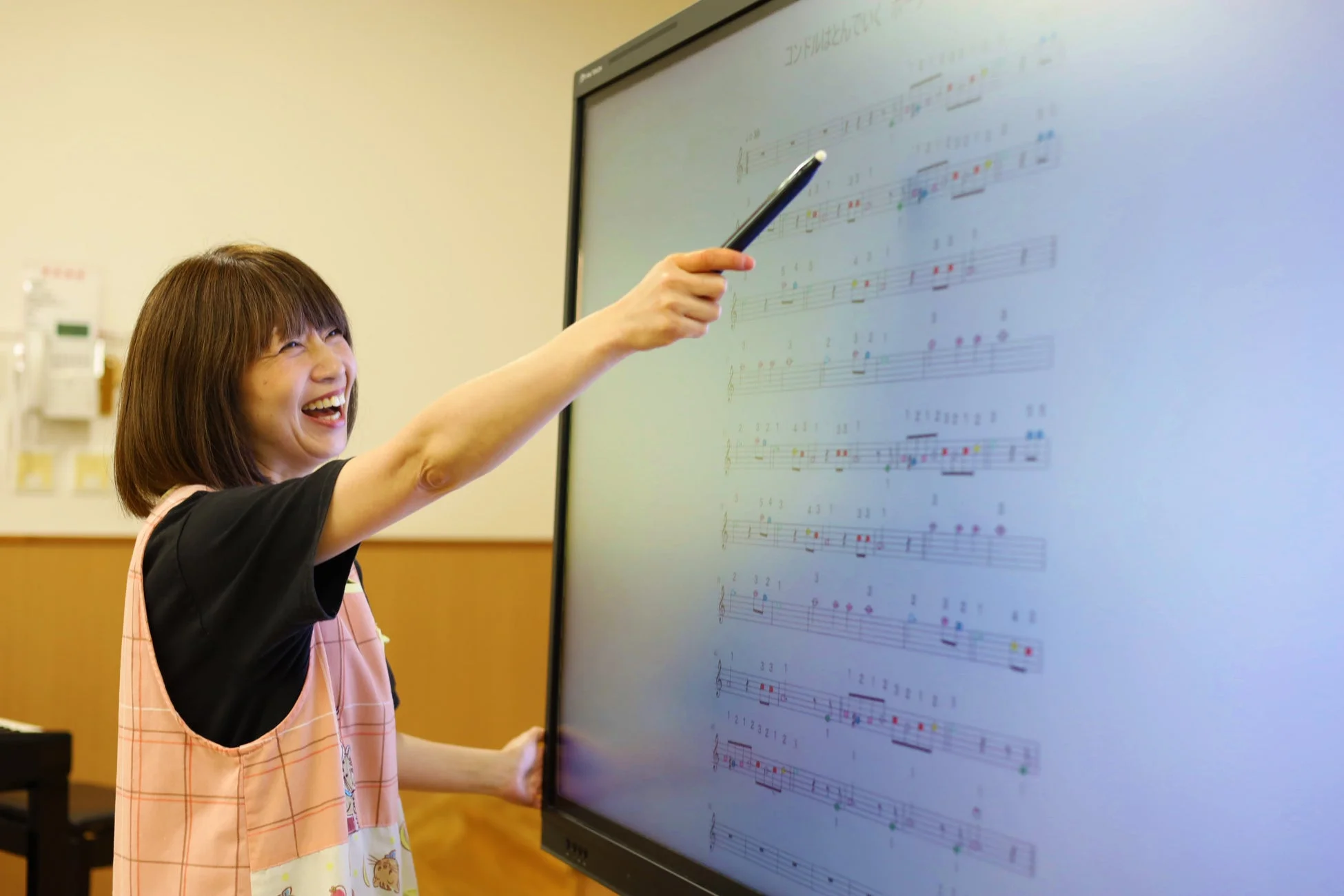 笑顔の女性がデジタル画面に表示された楽譜を指し示しています。画面には「コンドルはとんでいく」の楽譜が色付きで表示されており、音楽の授業や指導の様子を捉えた一枚です。