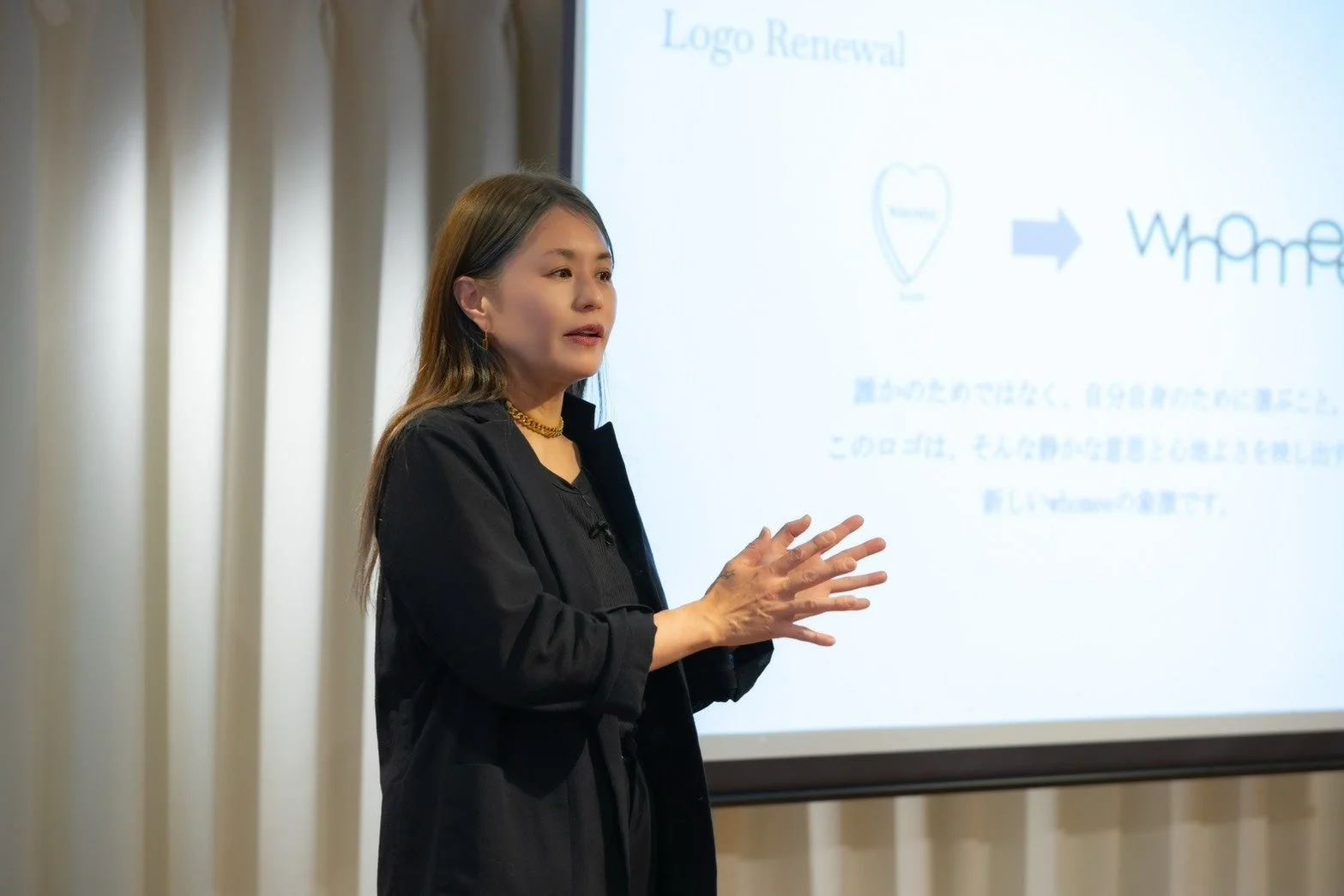女性がプレゼンテーションを行っており、スクリーンには「Logo Renewal」と書かれ、ブランド「Whomee」のロゴ変更について説明されています。