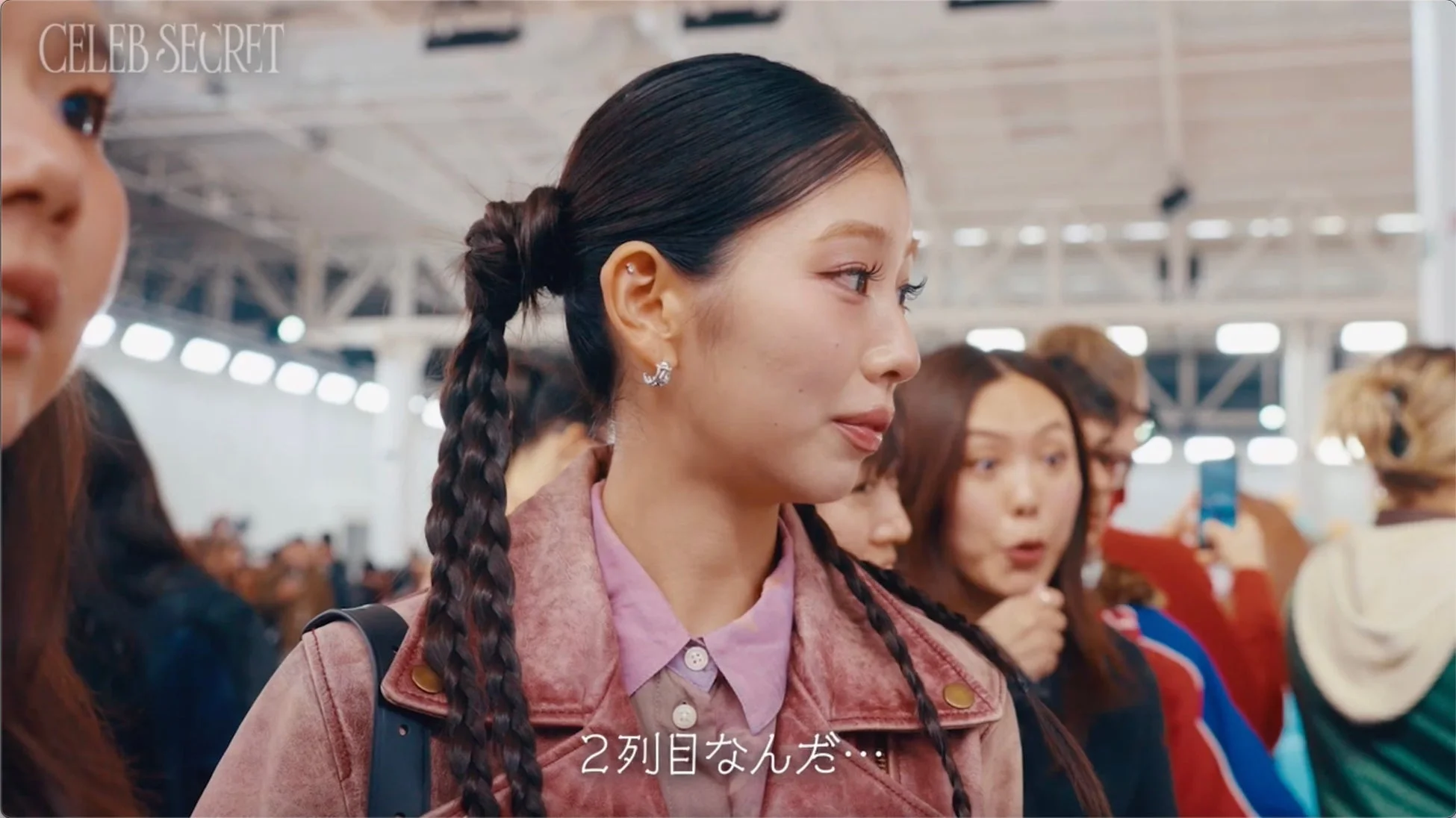 イベント会場のような場所で、編み込みのポニーテールとピンクのジャケットを着た女性が横を向いているクローズアップ画像です。周囲には他の人々が見え、「2列目なんだ…」というテキストが写っています。