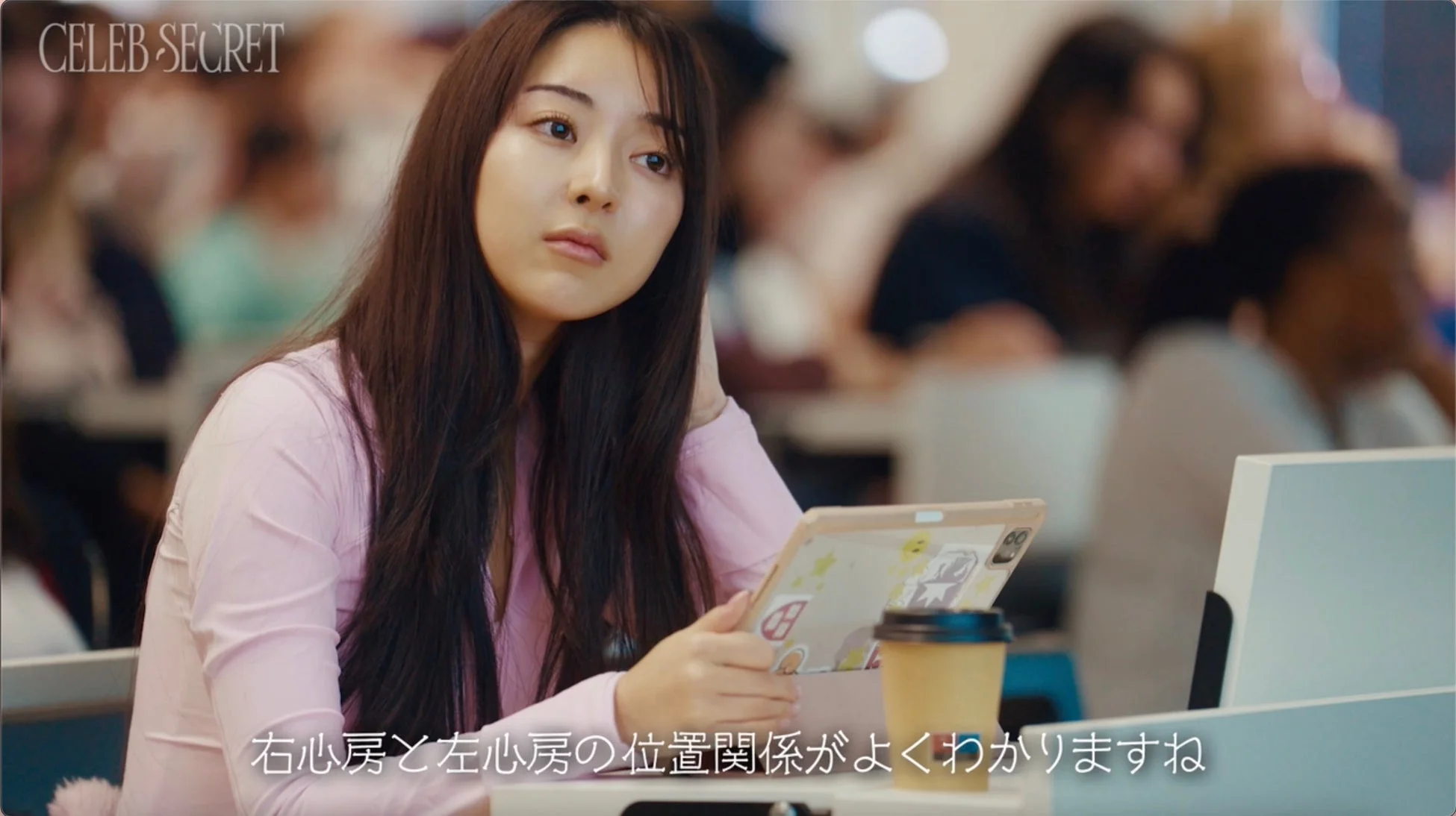 ピンクのトップスを着た若い女性が、タブレットを手に持ち、少し考え込んだような表情で座っています。カフェのような屋内空間で、背景には他の人々がぼやけて写っており、テーブルには飲み物も置かれています。画面下には「右心房と左心房の位置関係がよくわかりますね」というテキストが表示されています。