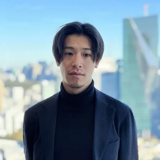 青い空と都市のビル群を背景に、黒いタートルネックとジャケットを着用したアジア人男性のポートレート