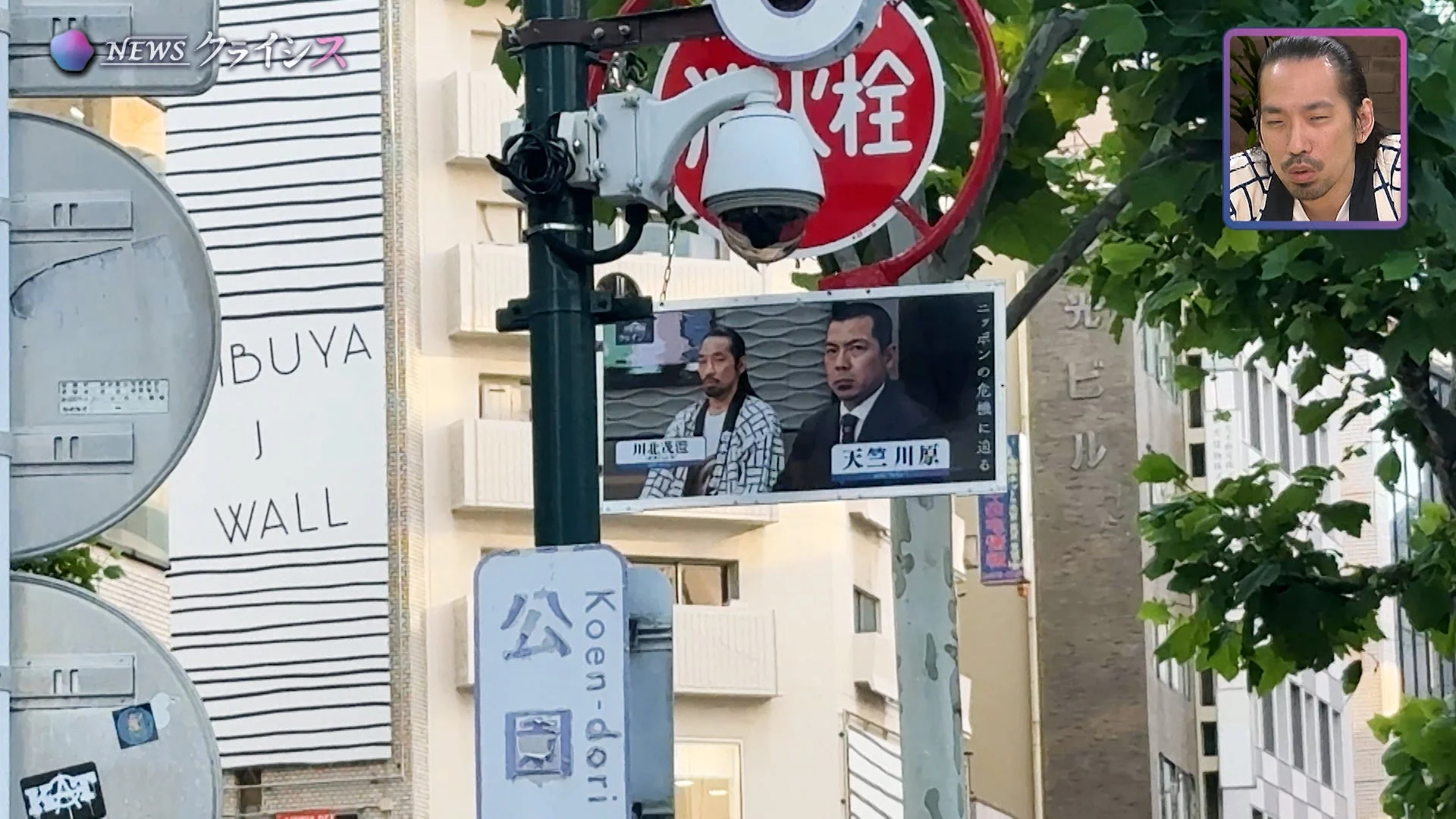 日本の都市の街角を捉えた画像。電柱には監視カメラや消火栓の標識があり、二人の男性が映る「ニッポンの危機に迫る」と書かれた看板が掲げられている。上部にはニュース番組のロゴと男性の顔が映る小窓が見える。公園通りの表示もある。