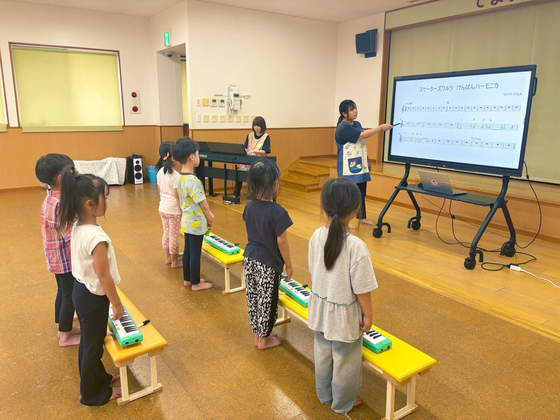 子供たちが教室で音楽の授業を受けている様子です。先生が大きなスクリーンに映し出された楽譜を指し示して説明しており、子供たちは鍵盤ハーモニカを前に真剣な表情で見ています。
