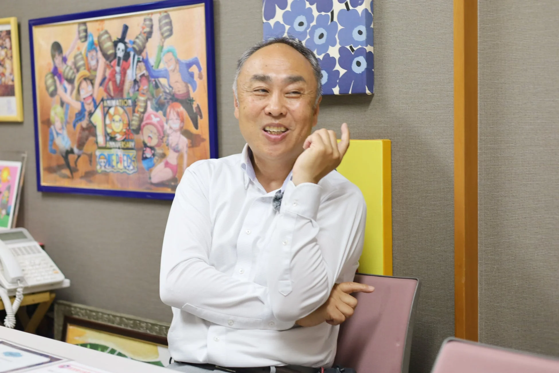 白いシャツを着た日本人男性が笑顔で座っており、背景には人気アニメ「ワンピース」の10周年記念ポスターが飾られています。