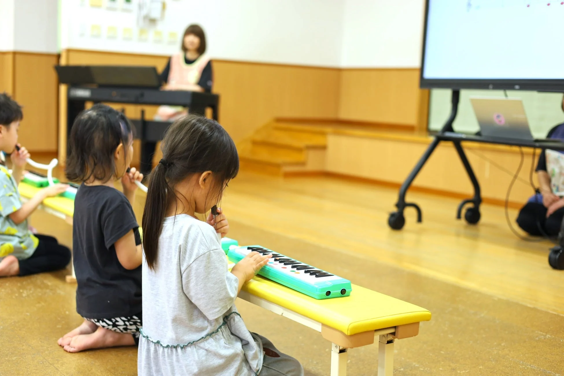 複数の子供たちが音楽教室で鍵盤ハーモニカを演奏している様子です。先生がピアノを弾き、大きなスクリーンには楽譜が映し出されており、楽しく音楽を学んでいます。