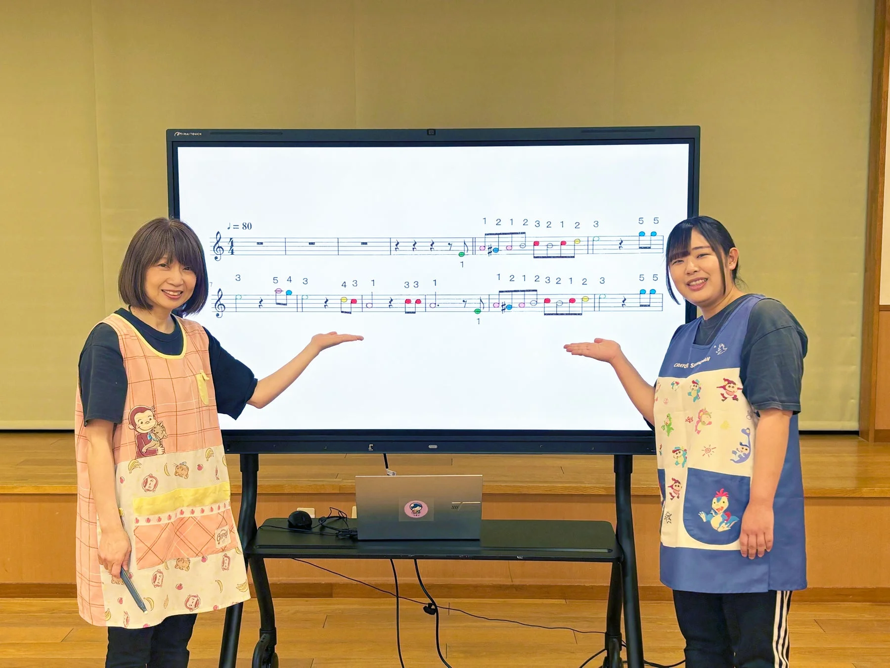 音楽の楽譜が映し出された電子黒板の前で、エプロンをつけた二人の女性が笑顔で指導している様子